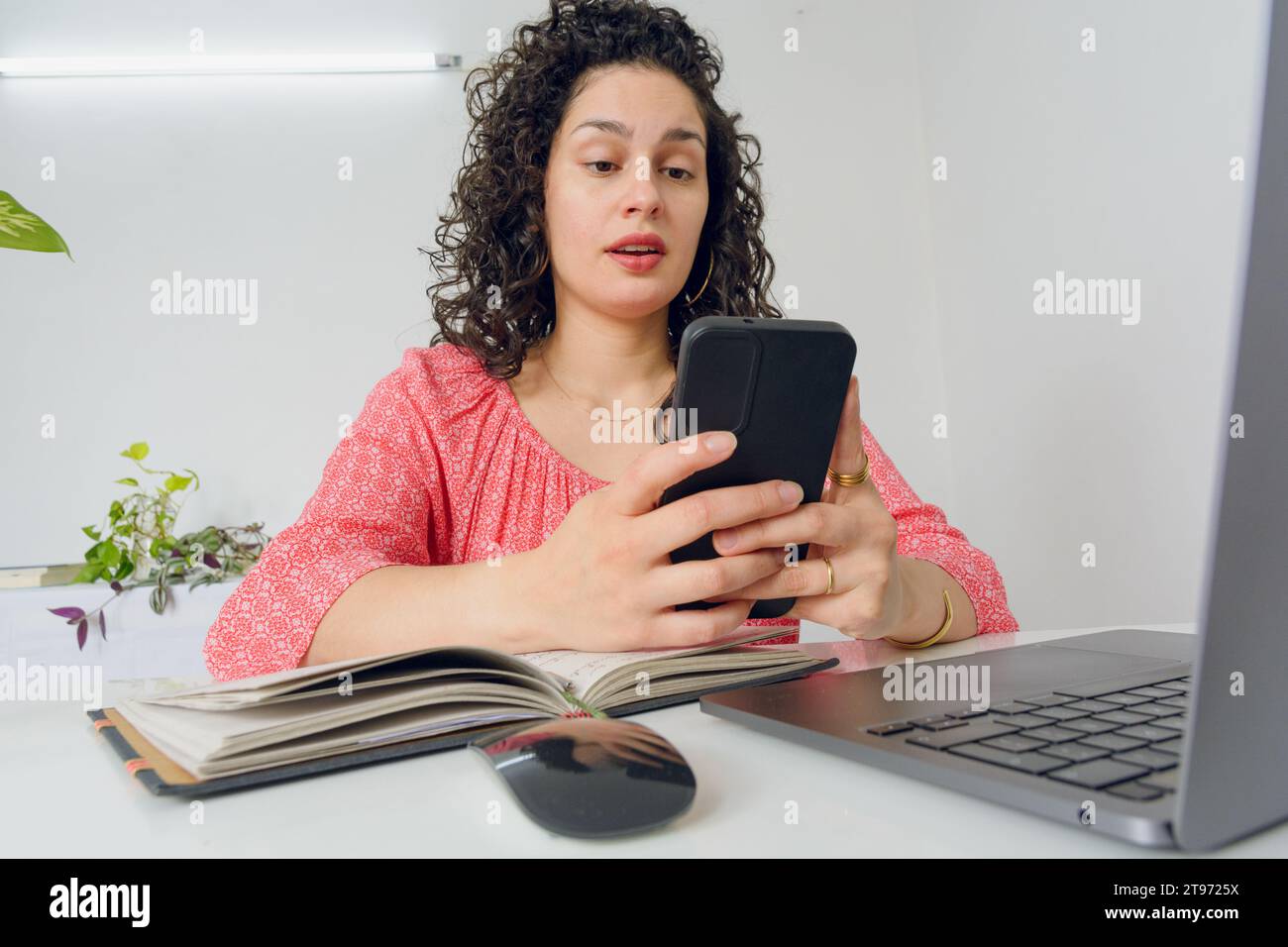 Jeune femme vénézuélienne latine, travailleur dans son bureau à domicile assis avec son ordinateur portable surpris avec sa bouche ouverte en utilisant le téléphone lisant des messages et social Banque D'Images
