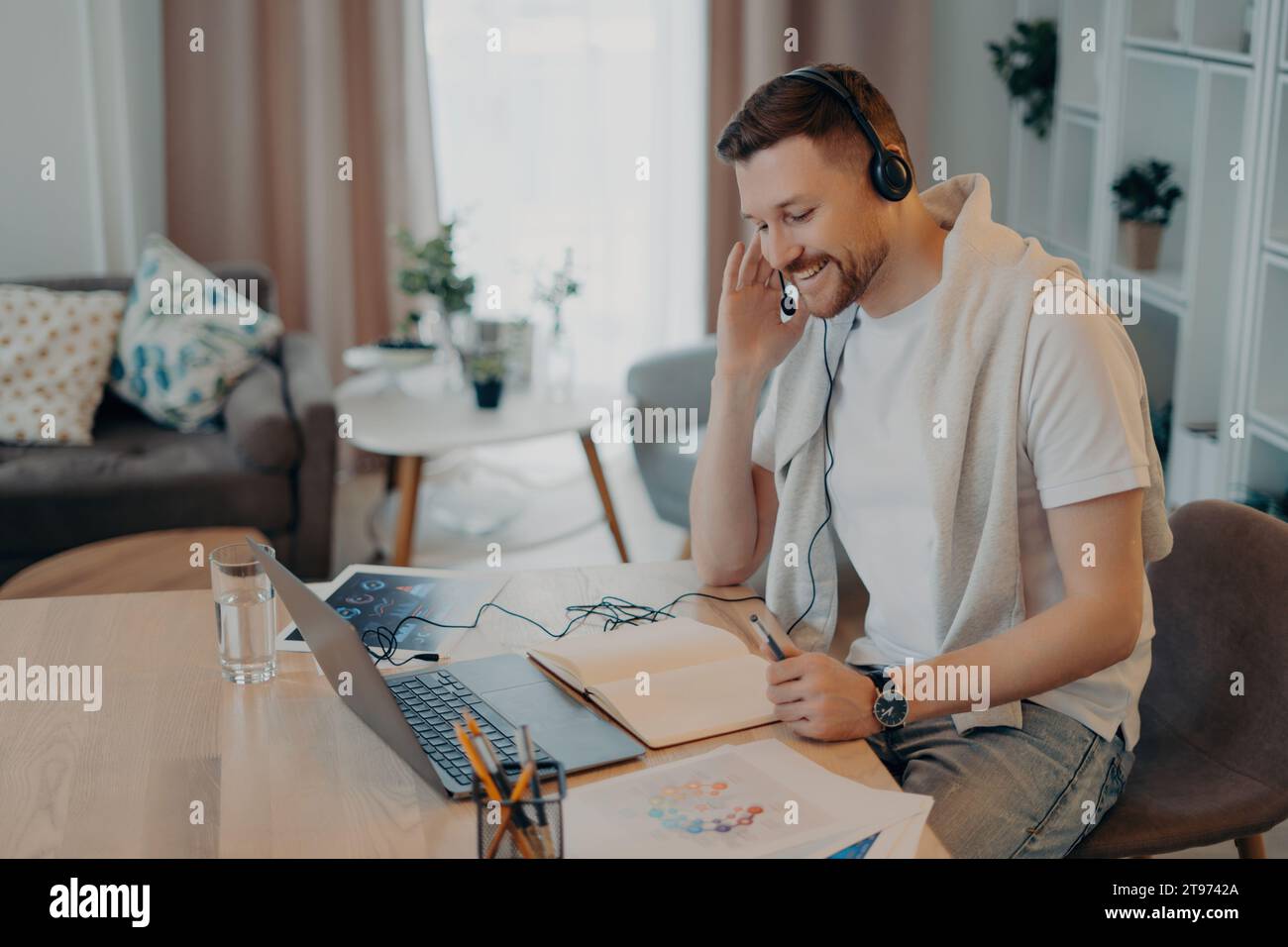 Freelance joyeuse avec des écouteurs engagés dans un appel en ligne dans une configuration confortable à la maison, illustrant le mode de vie moderne du télétravail Banque D'Images