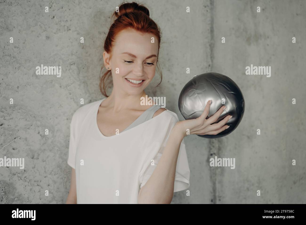 Joyeuse rousse dans une tenue décontractée pose de manière ludique avec une balle d'exercice argentée dans une salle de gym minimaliste Banque D'Images