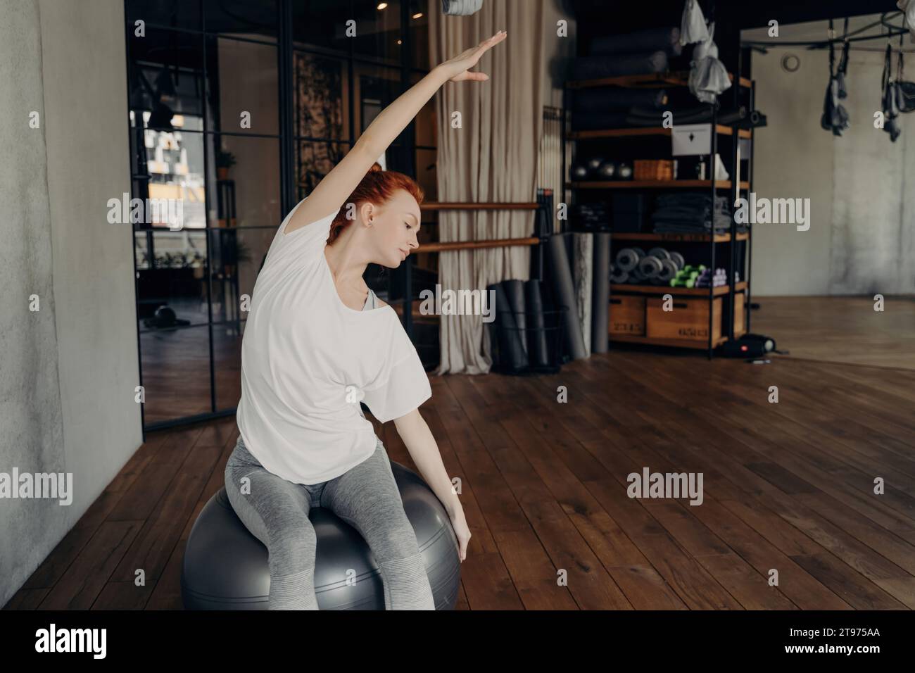 Femme rousse concentrée s'exerçant sur un ballon de fitness dans une salle de gym moderne, démontrant un étirement latéral pour la flexibilité Banque D'Images