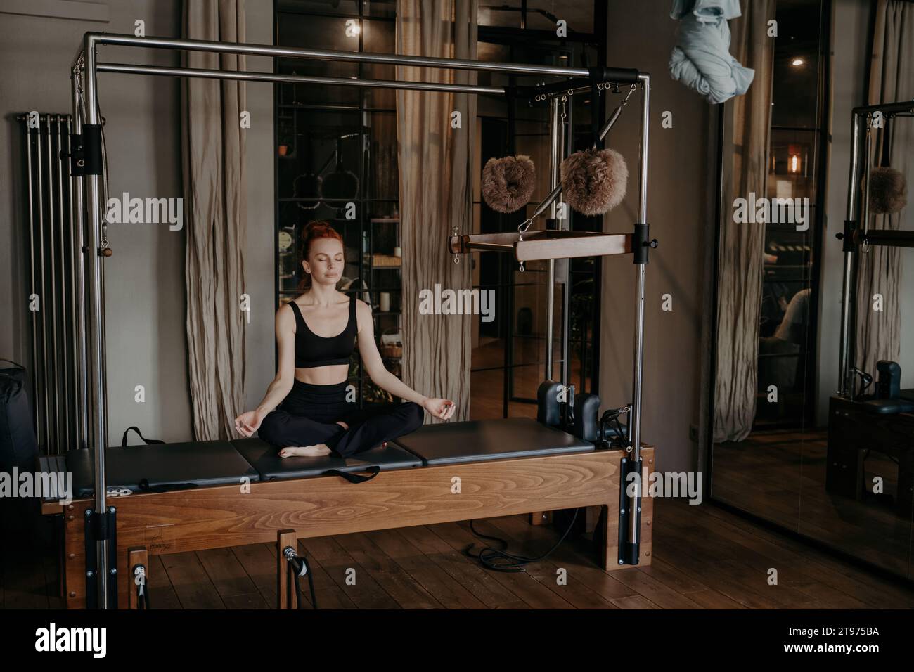 Sérénité en mouvement : une femme rousse en tenue noire médite sur un reformeur Pilates dans un studio tranquille Banque D'Images