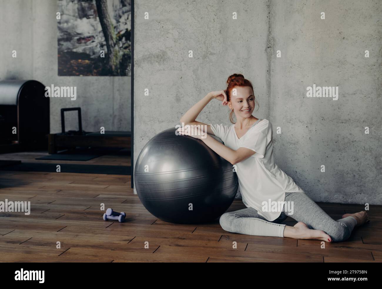 Redhead Femme en sourires de sportswear décontracté, se détendant à côté d'un ballon de fitness dans un studio d'entraînement ensoleillé Banque D'Images