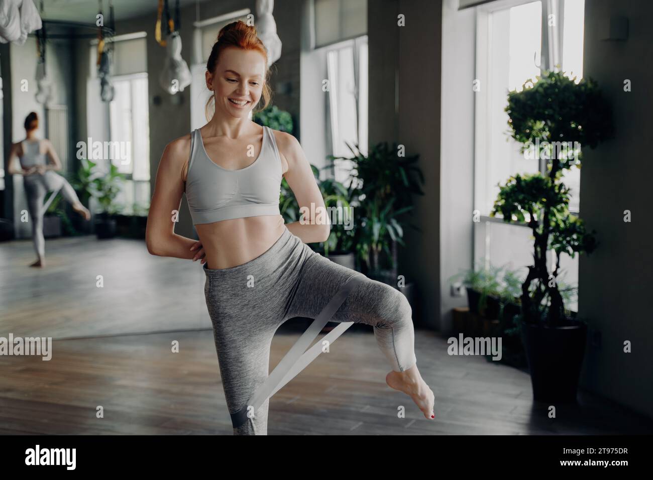 Joyeuse femme rousse en tenue de gym pratique gracieusement des exercices de bande de résistance dans un espace d'entraînement ensoleillé et rempli de plantes Banque D'Images