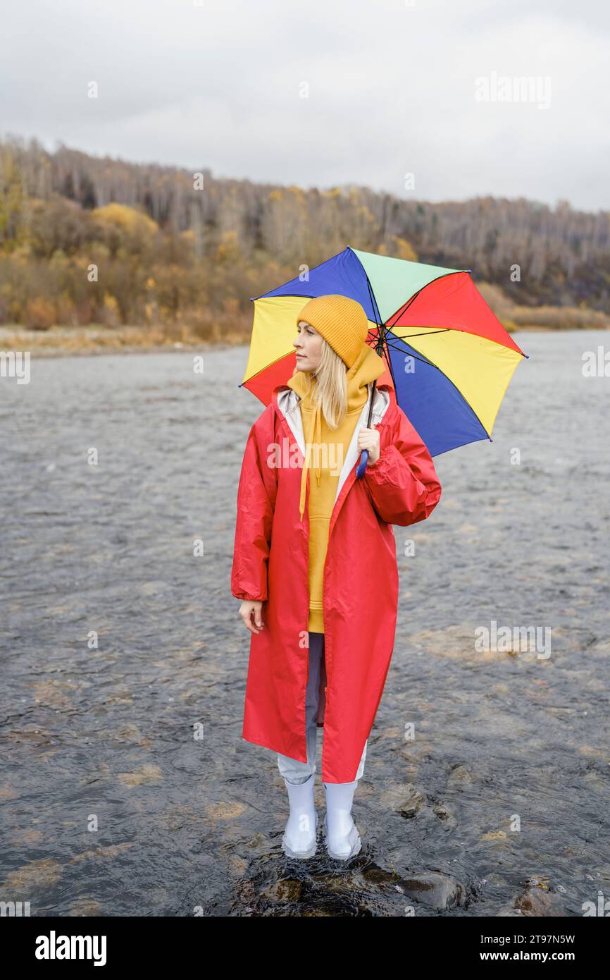 Femme contemplative portant un imperméable rouge debout avec un parapluie dans l'eau de la rivière Banque D'Images