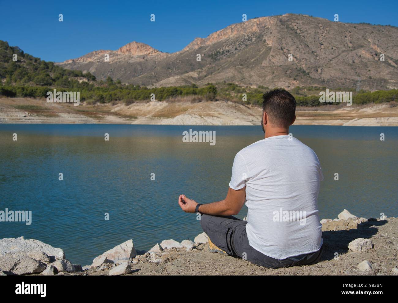 Homme adulte dans une pose détendue méditant au bord d'un lac par une journée ensoleillée. Idéal pour les projets liés aux sensations et aux émotions, au calme et aux sports de plein air. Banque D'Images