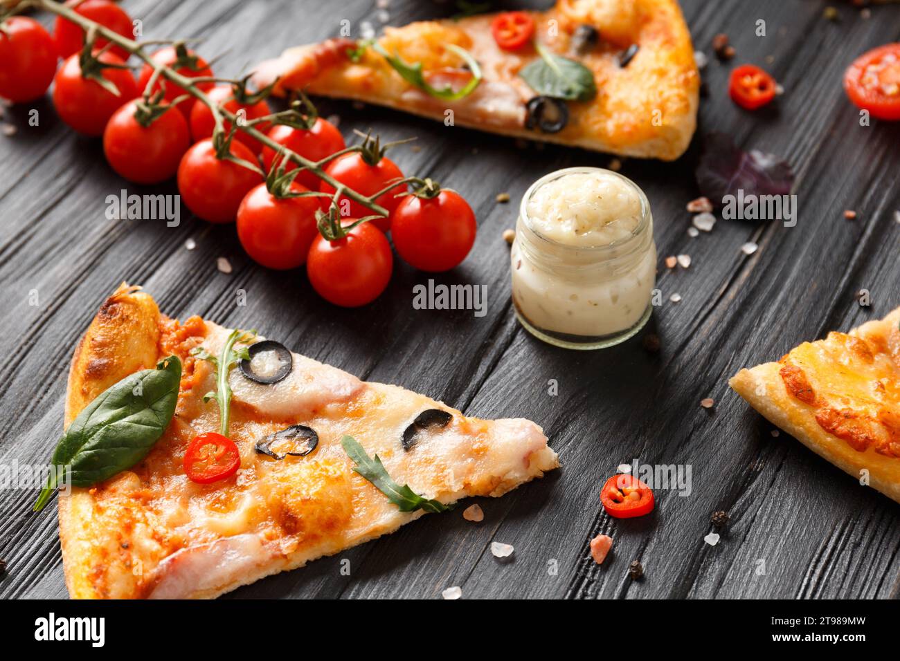 Des tranches de pizza avec jerky, des olives, du fromage et des herbes sont disposées sur un fond en bois foncé, une branche de tomates cerises fraîches, des sauces et des assaisonnements f Banque D'Images