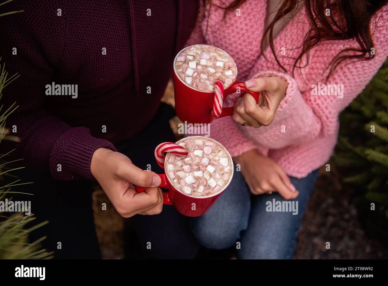 Gros plan des mains d'un jeune couple tenant des tasses rouges avec du cacao chaud, des guimauves, du rouge rayé, de la canne à sucre blanche. L'homme et la femme s'amusent à Christ Banque D'Images