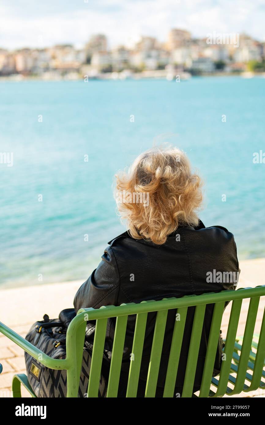 Vieillesse - seul. Vieille femme avec sac vu de derrière assis regardant vers la mer. Grand-mère solitude. Le retraité est assis sur un banc, Banque D'Images