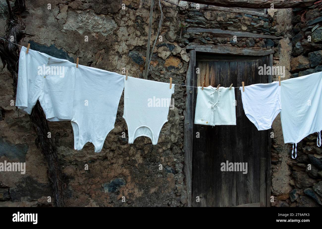 Une corde à linge maison avec des vêtements vintage blancs suspendus, en arrière-plan la façade d'une ancienne maison canarienne, avec une porte en bois et une pierre W. Banque D'Images