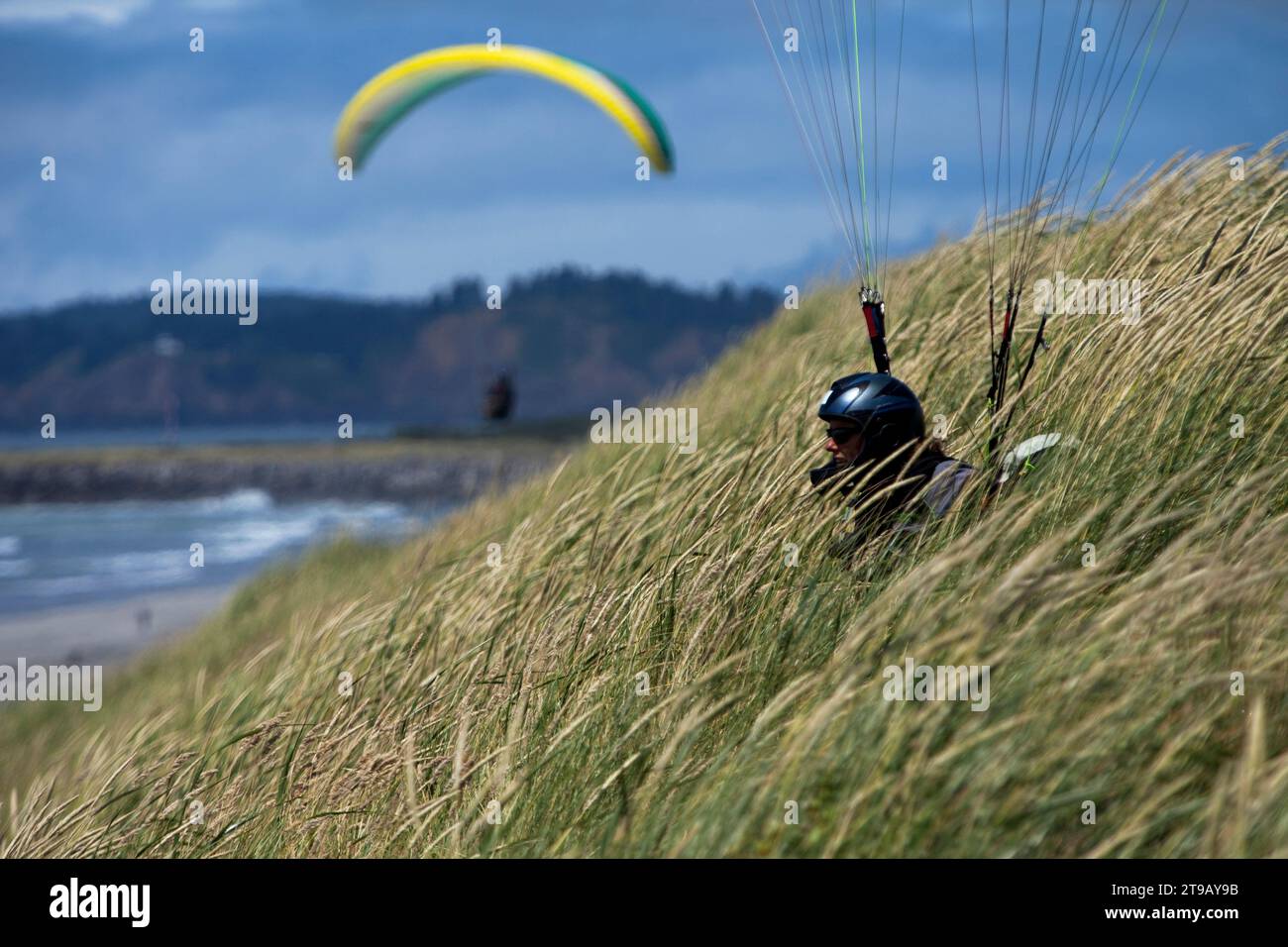 Parapente féminine assise dans de hautes herbes sur une dune au-dessus d'une plage tandis qu'un autre parapente vole en arrière-plan. Banque D'Images