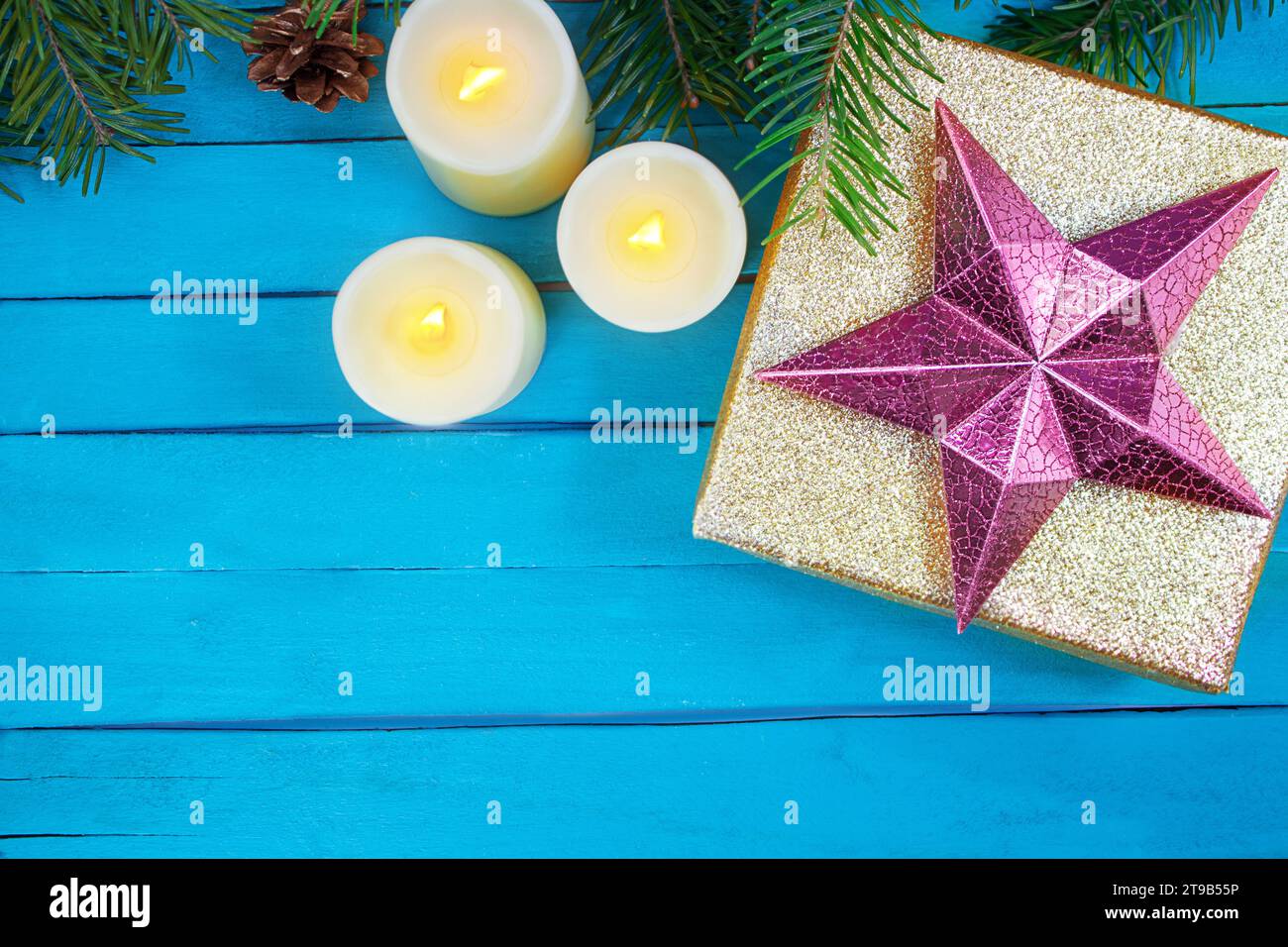 Boîte brillante dorée avec étoile rose, bougies allumées avec feu et pomme de pin, branches de sapin sur des planches bleues. Noël, nouvel an, anniversaire, vacances. Copier sp Banque D'Images