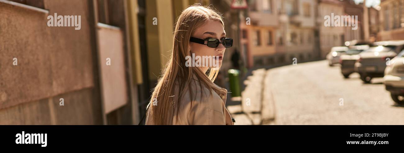 jeune femme blonde en trench élégant et lunettes de soleil marchant dans la rue en journée ensoleillée, bannière Banque D'Images