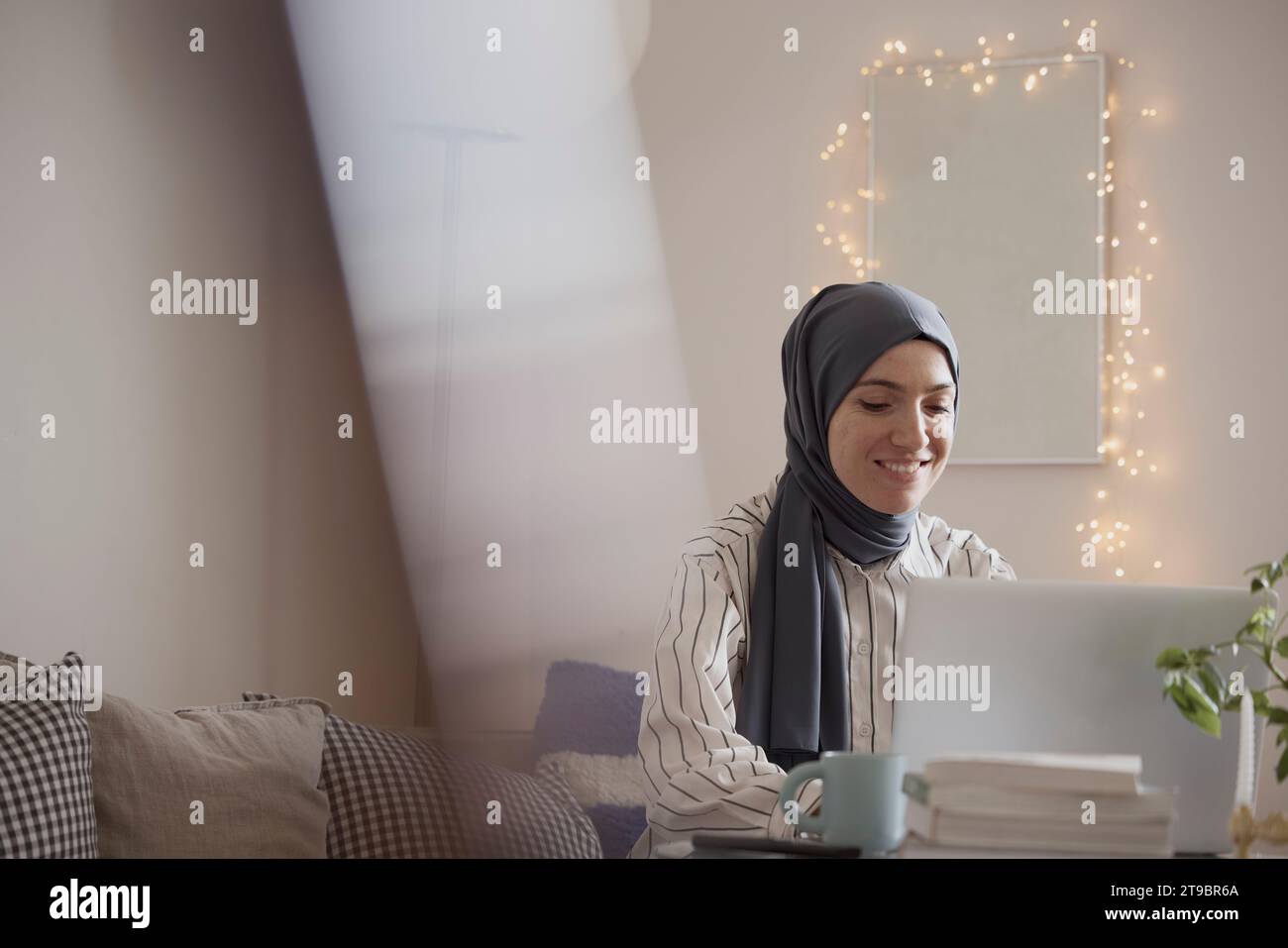 Femme pigiste souriante portant le hijab tout en travaillant sur ordinateur portable dans le salon à la maison Banque D'Images