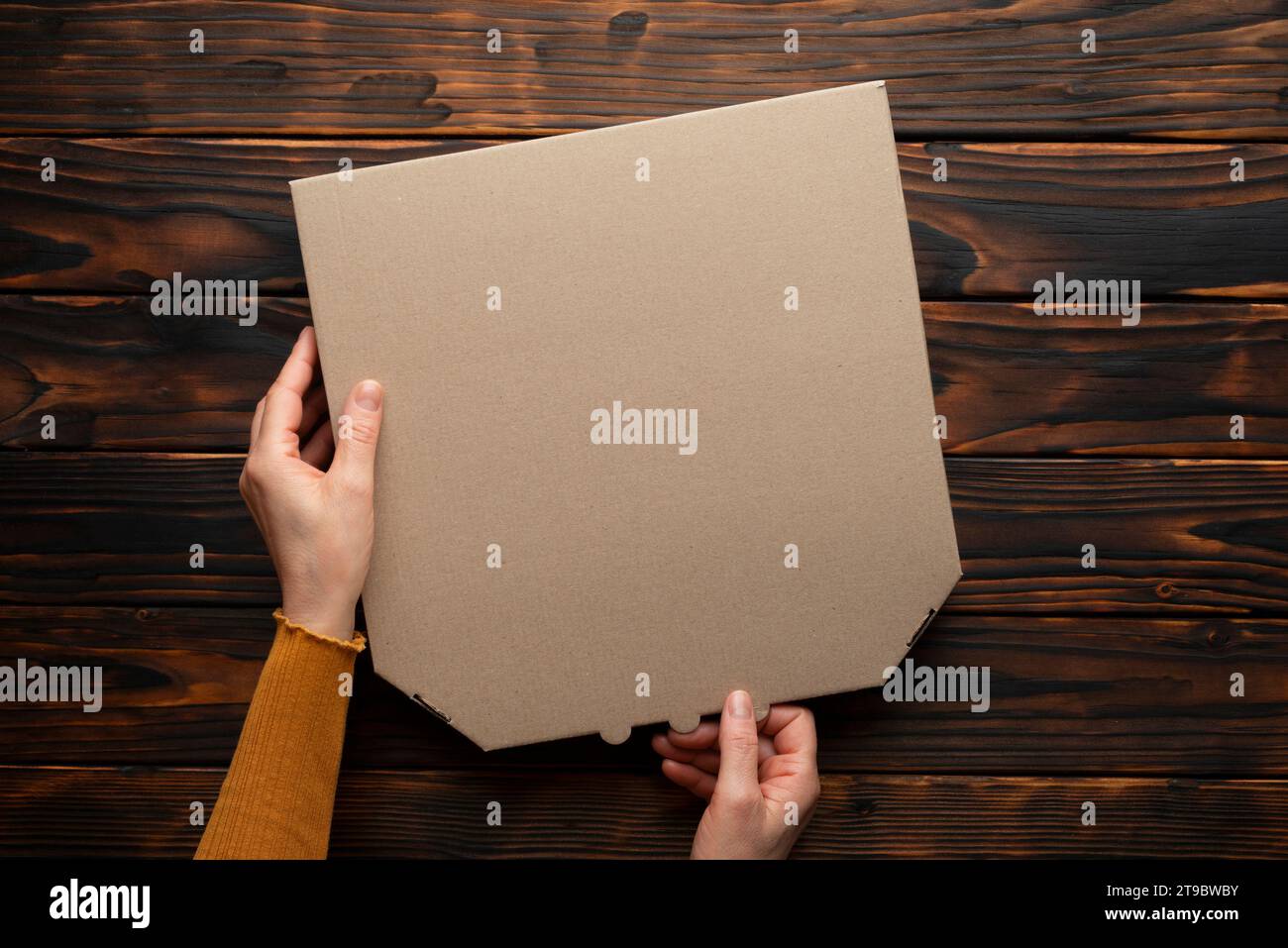 Boîte de carton fermée de pizza dans les mains caucasiennes sur la maquette plate de table en bois foncé naturel avec l'espace de copie Banque D'Images