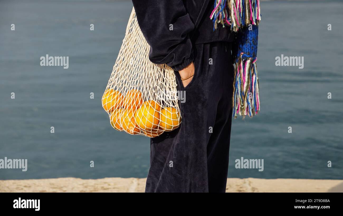 Fille en costume de velours bleu marine tenant des mandarines orange vif dans un sac à cordes beige. Banque D'Images