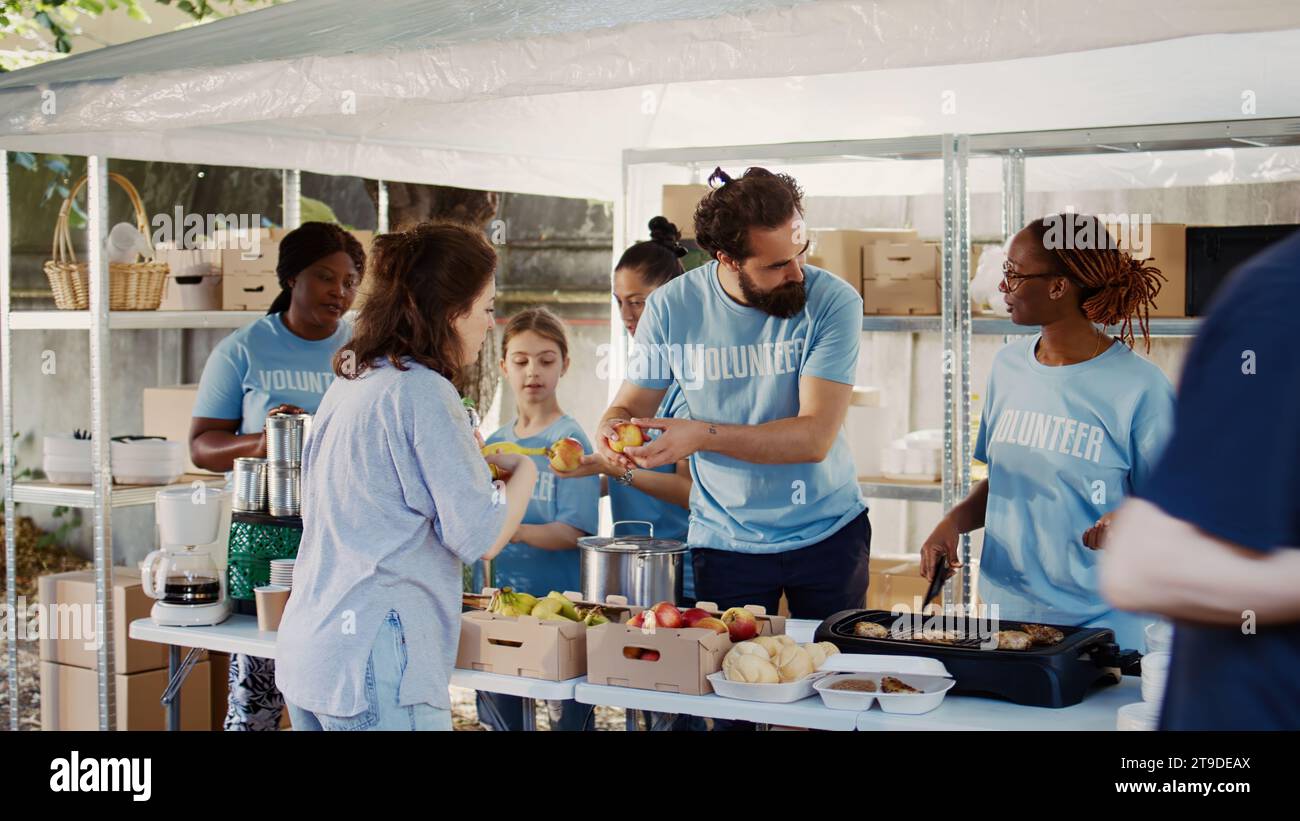 Des personnes volontaires portant des t-shirts bleus servant gratuitement de la nourriture fraîchement préparée et des provisions aux personnes confrontées à la faim et à la pauvreté. Hommes et femmes travailleurs caritatifs distribuant des repas chauds aux nécessiteux. Banque D'Images