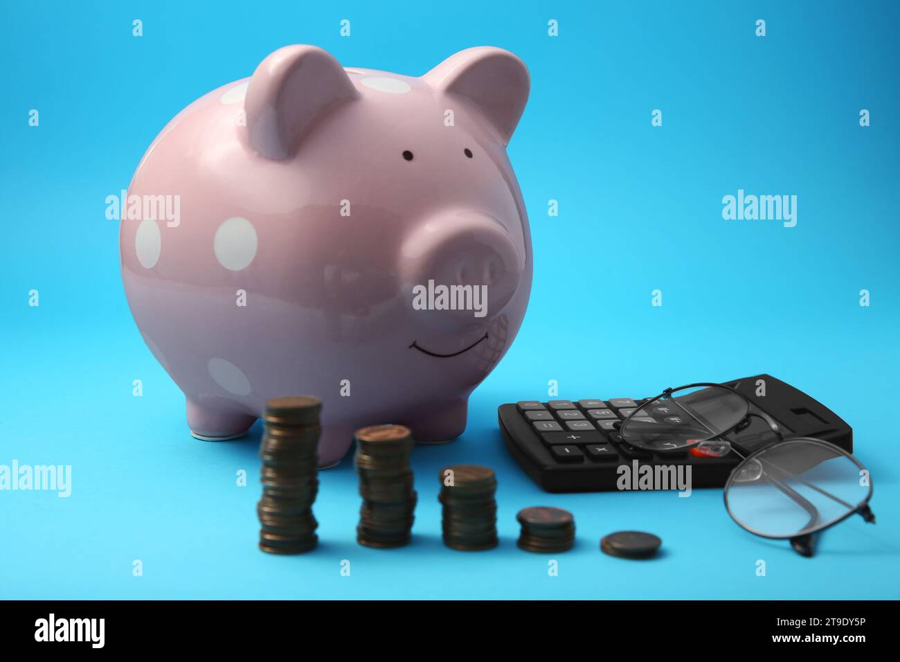 Tirelire, piles de pièces de monnaie, lunettes et calculatrice sur fond bleu clair Banque D'Images