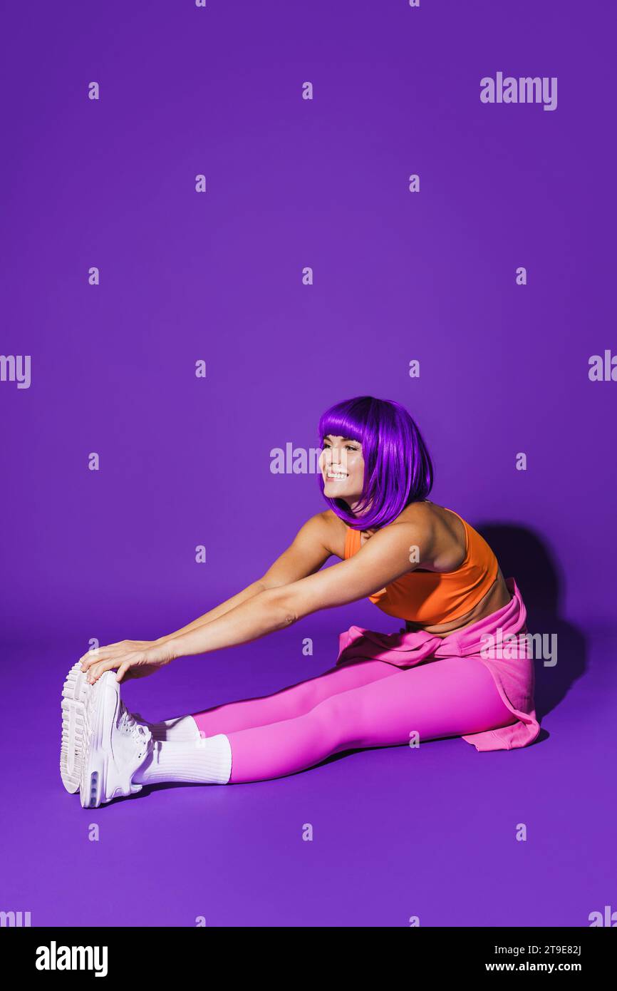Jeune femme joyeuse portant des vêtements de sport colorés pendant l'entraînement d'étirement sur fond violet Banque D'Images