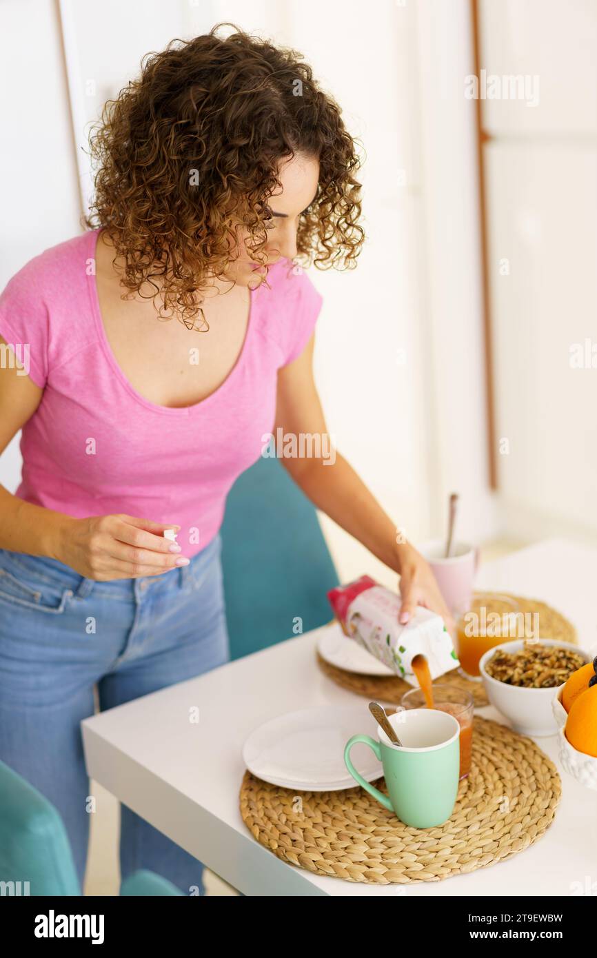 Femme adulte dans des vêtements décontractés et avec les cheveux bouclés regardant vers le bas tout en se tenant près de la table avec le verre, sur le napperon et en versant le jus de fruit du paquet Banque D'Images