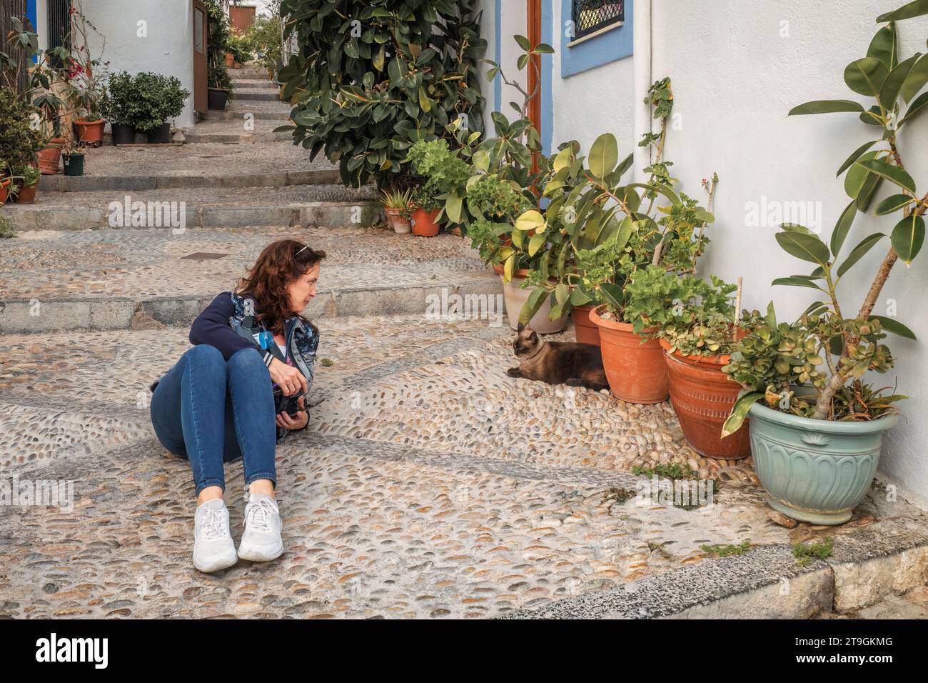 Femme couchée sur le sol portant un Jean et une veste parlant à un chat dans une rue pavée avec des escaliers dans la ville de Peñiscola, Castellon, Espagne. Banque D'Images