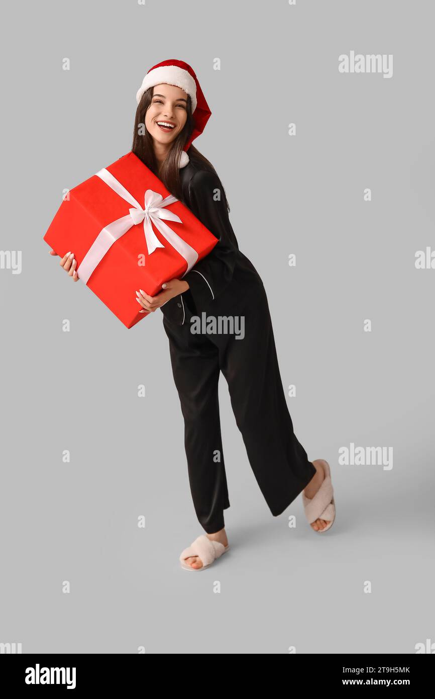 Belle jeune femme en pyjama noir et chapeau de Père Noël avec boîte cadeau de Noël sur fond gris Banque D'Images