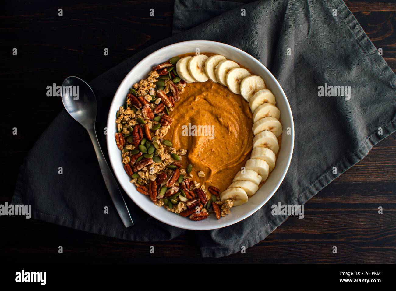 Bol de smoothie à la citrouille sur une table en bois avec une cuillère : smoothie à la citrouille garni de granola, de pacanes, de graines de citrouille et de bananes tranchées Banque D'Images