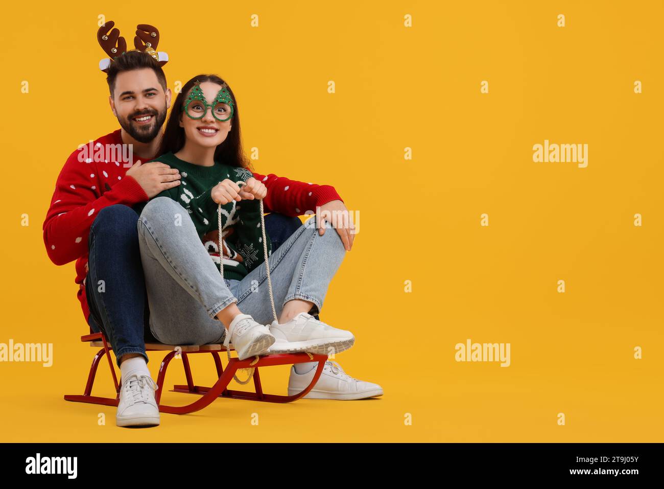 Heureux jeune couple dans des pulls de Noël, bandeau de renne et lunettes de fête sur le traîneau sur fond orange. Espace pour le texte Banque D'Images