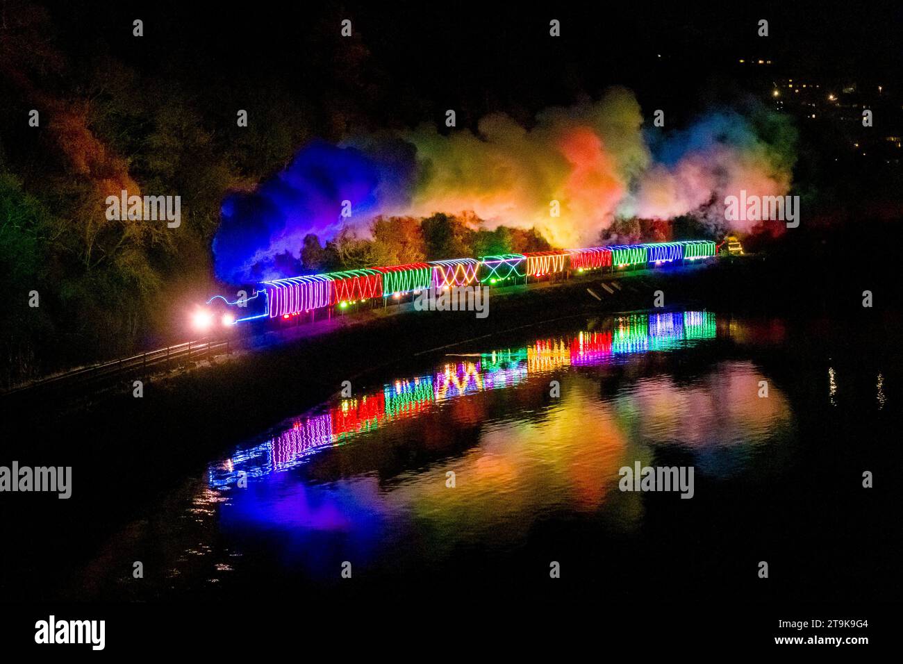 Kingswear, Devon, Royaume-Uni. 25 novembre 2023. Le train des lumières de Noël sur le chemin de fer à vapeur de Dartmouth dans le Devon, qui fonctionne jusqu'au 30 décembre 2023, se reflète dans les eaux calmes de la rivière Dart après avoir quitté Kingswear pour son voyage de retour à Paignton. Les voitures et les locomotives à vapeur du service festif sont éclairées à l'intérieur et à l'extérieur avec des lumières colorées. Crédit photo : Graham Hunt/Alamy Live News Banque D'Images