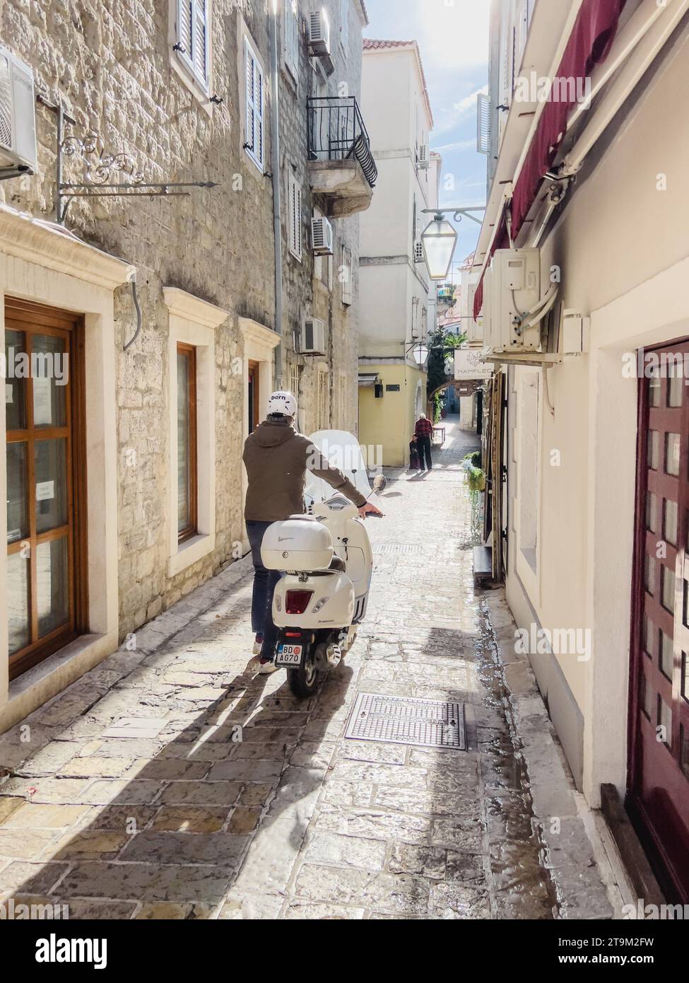 Budva, Monténégro - 17 août 2023 : un homme portant un casque conduit un scooter dans ses mains le long d'une rue étroite en pierre d'une ville antique Banque D'Images