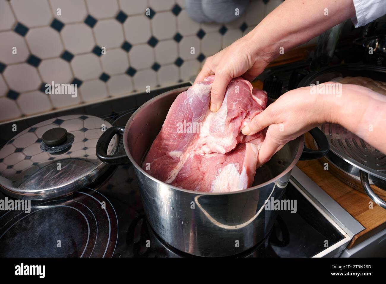 Mains de femme plaçant de la viande de porc crue dans une casserole en acier inoxydable à bouillir sur le dessus de la cuisinière dans la cuisine, préparation de cuisson pour un dîner de fête, Banque D'Images