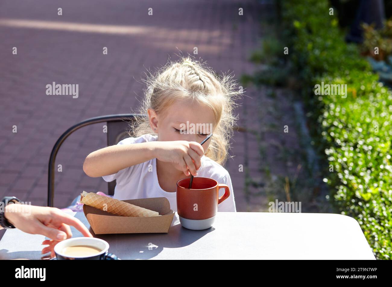 Petite fille souriante avec la mère prendre un petit déjeuner dans le café d'été en plein air. Mignonne fille caucasienne mangeant un tube à gaufres savoureux et buvant du cacao Banque D'Images