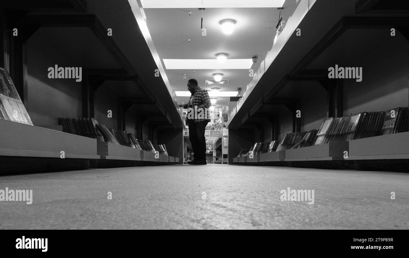 Homme noir d'âge moyen au bout d'une allée dans un magasin de disques regardant à travers les piles ; magasiner de la musique ; image monochrome en noir et blanc. Banque D'Images