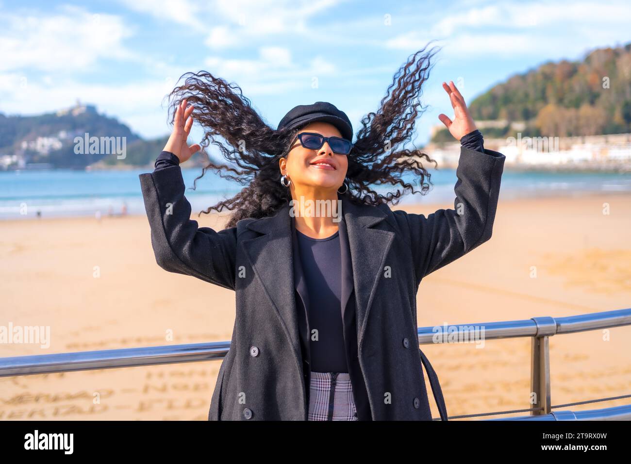 Chic femme latine dans des vêtements d'hiver arrangeant ses cheveux dans un point de vue de plage Banque D'Images