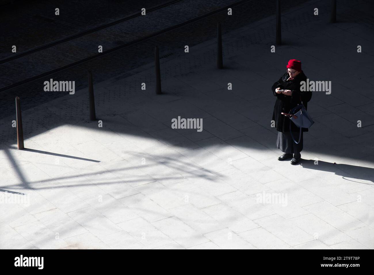 Belgrade, Serbie- 18 mars 2022 : une femme âgée avec un chapeau rouge debout seule sur une rue de la place de la ville par la journée froide et ensoleillée, vue à grand angle Banque D'Images