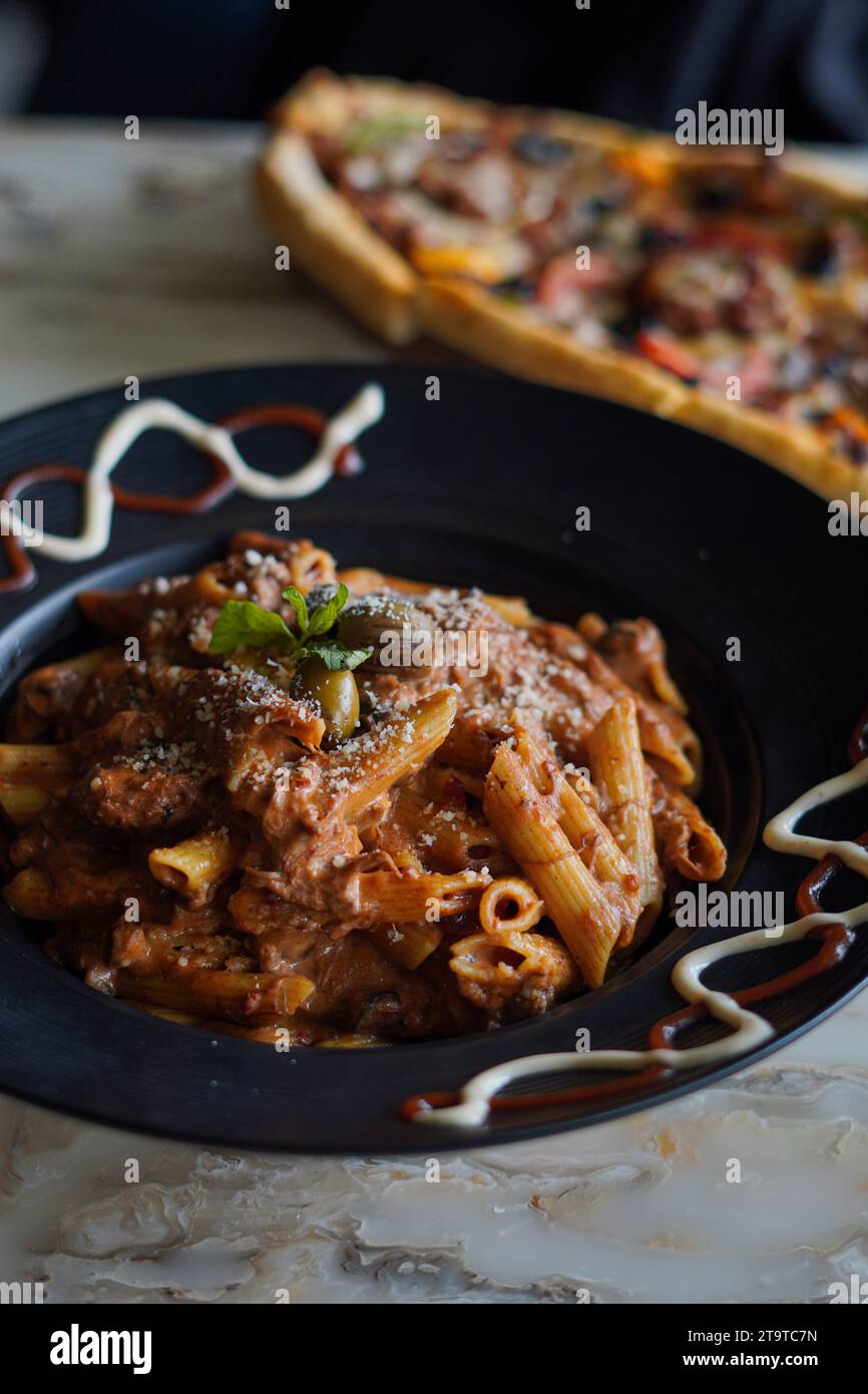 Pâtes Penne avec sauce, olive et fromage parmesan fraîchement râpé, dans une assiette noire, avec sauce sur le côté. Et une pizza fraîche en arrière-plan. Banque D'Images