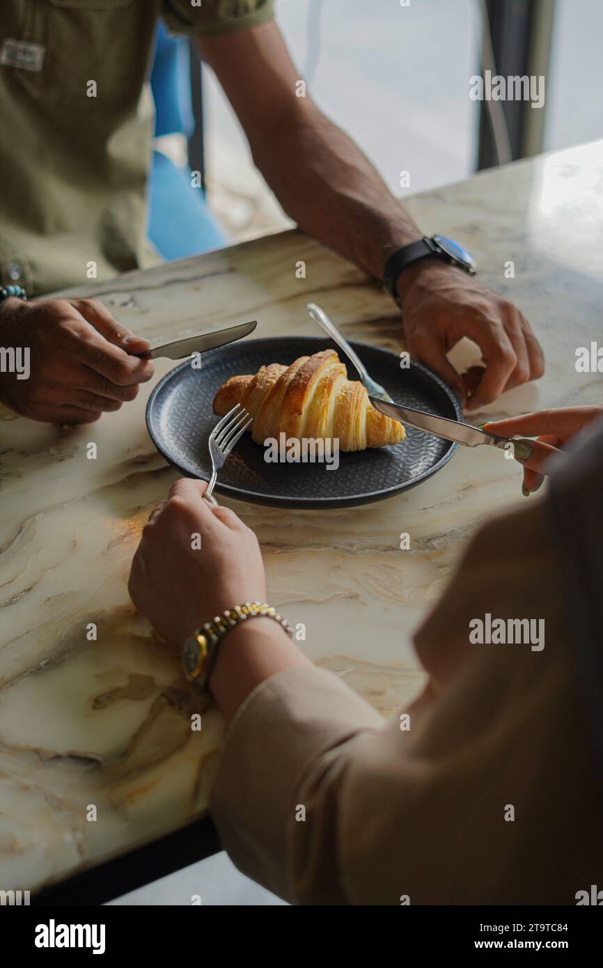 Un croissant sur assiette noire, deux personnes voulant le couper avec un couteau et une fourchette. Banque D'Images