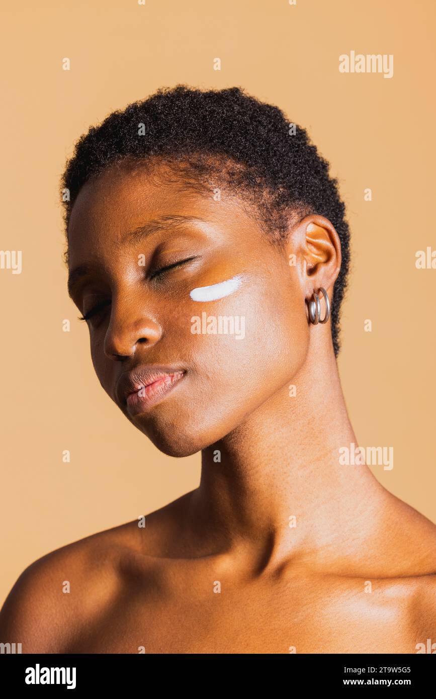 Jeune femme noire les yeux fermés avec une crème hydratante sur le visage. Jeune femme traitant sa peau du visage avec un produit de beauté. Banque D'Images