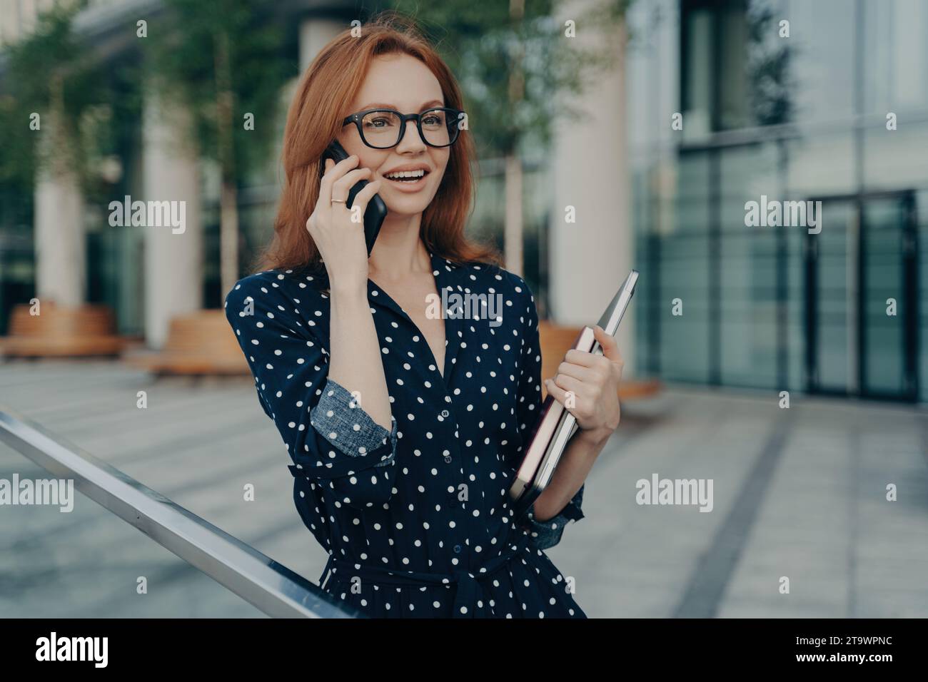 Femme d'affaires souriante en pois faire un appel téléphonique tout en tenant un ordinateur portable, à l'extérieur Banque D'Images