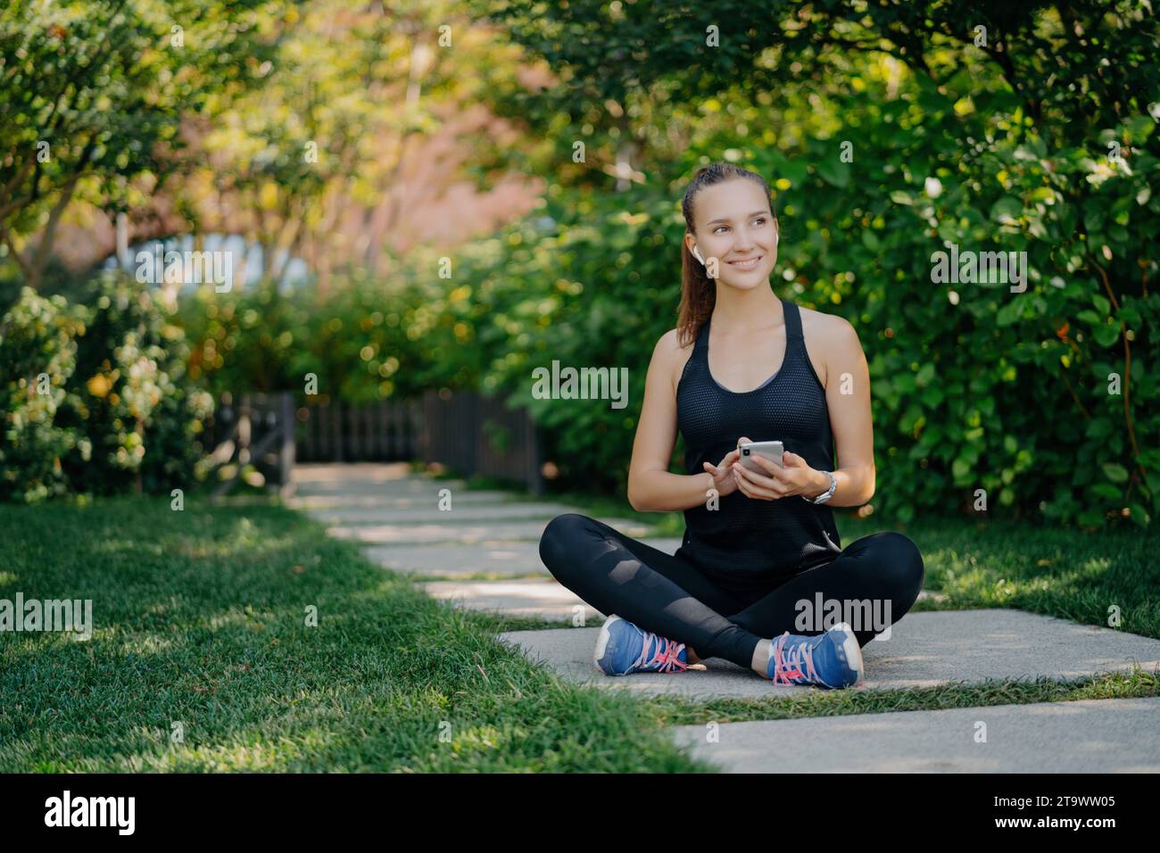 Heureuse jeune femme assise les jambes croisées sur un chemin dans un parc, tenant un smartphone, portant des vêtements de sport Banque D'Images