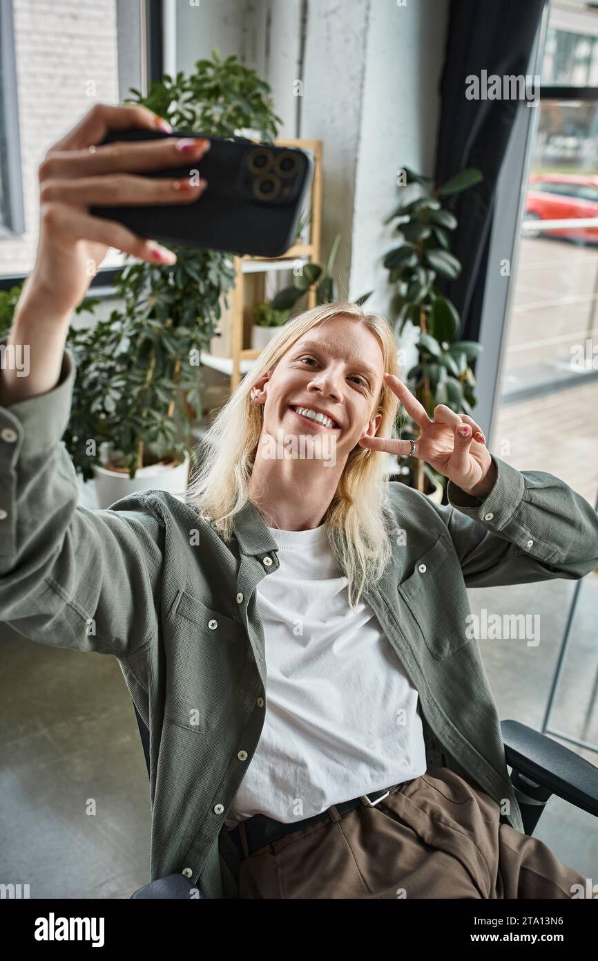 plan vertical d'une personne androgyne aux cheveux longs et joyeux en tenue décontractée prenant un selfie au bureau Banque D'Images