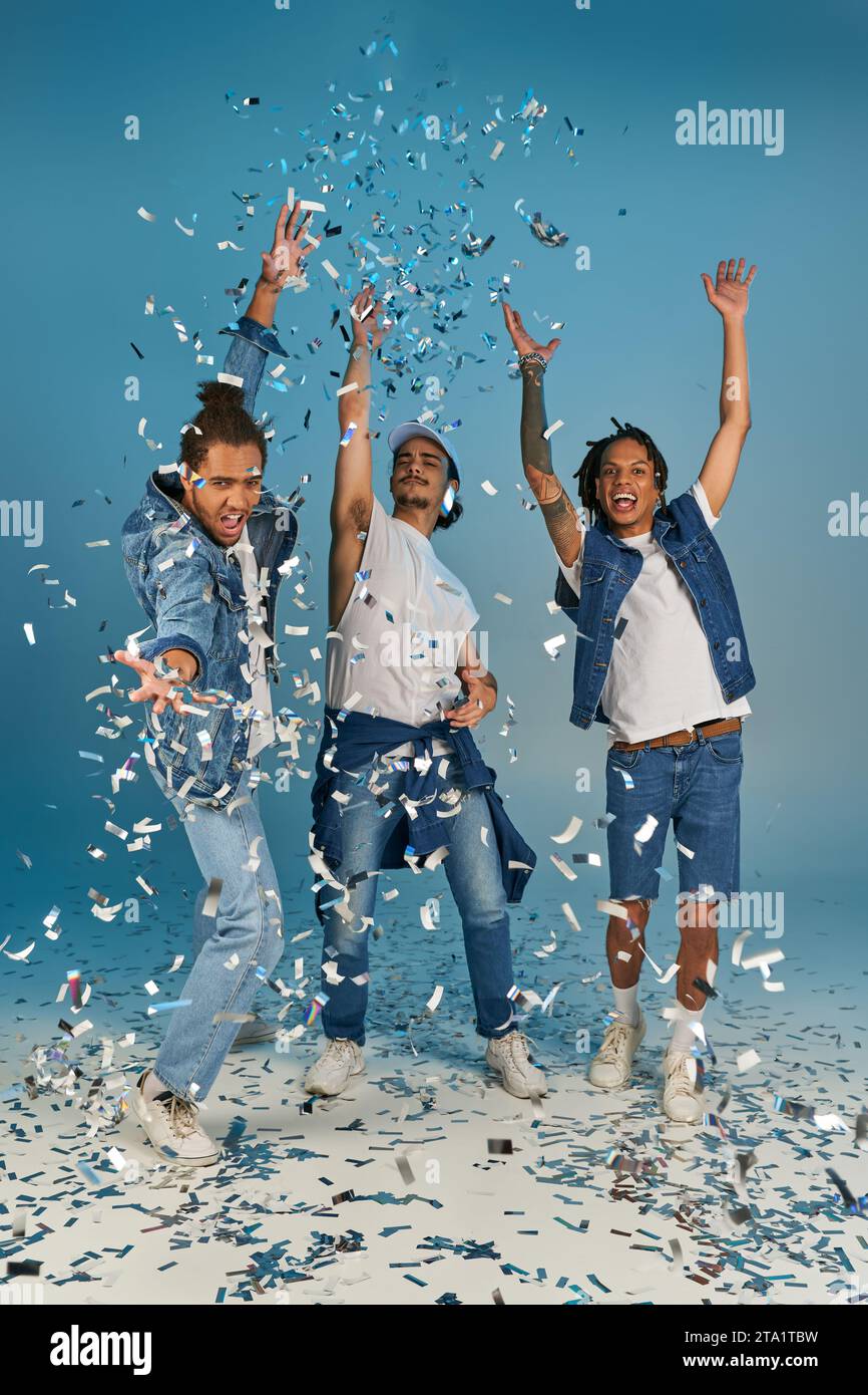 hommes multiculturels excités dans des tenues de denim à la mode se réjouissant sous la pluie brillante de confettis sur le bleu Banque D'Images