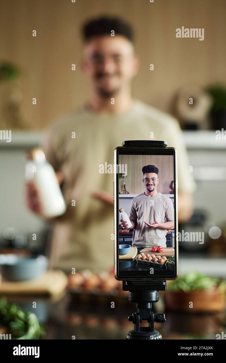 Gros plan sur smartphone filmant un jeune chef du Moyen-Orient souriant montrant une bouteille de lait à la caméra tout en se tenant près de la table de cuisine, concentrez-vous sur le smartphone Banque D'Images