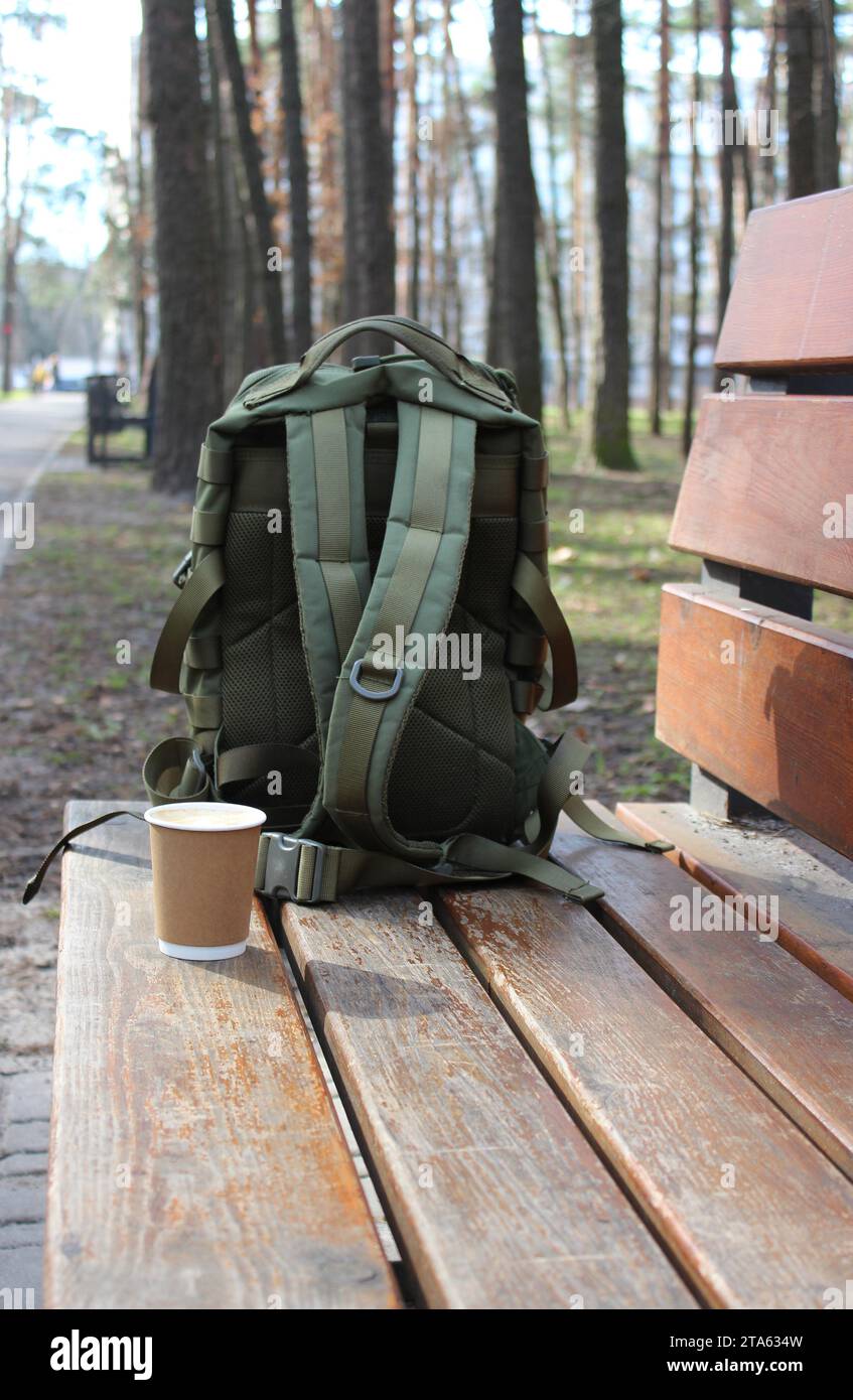 Un sac de voyage avec des sangles recouvre une tasse de papier de café du vent sur un banc dans le parc Banque D'Images