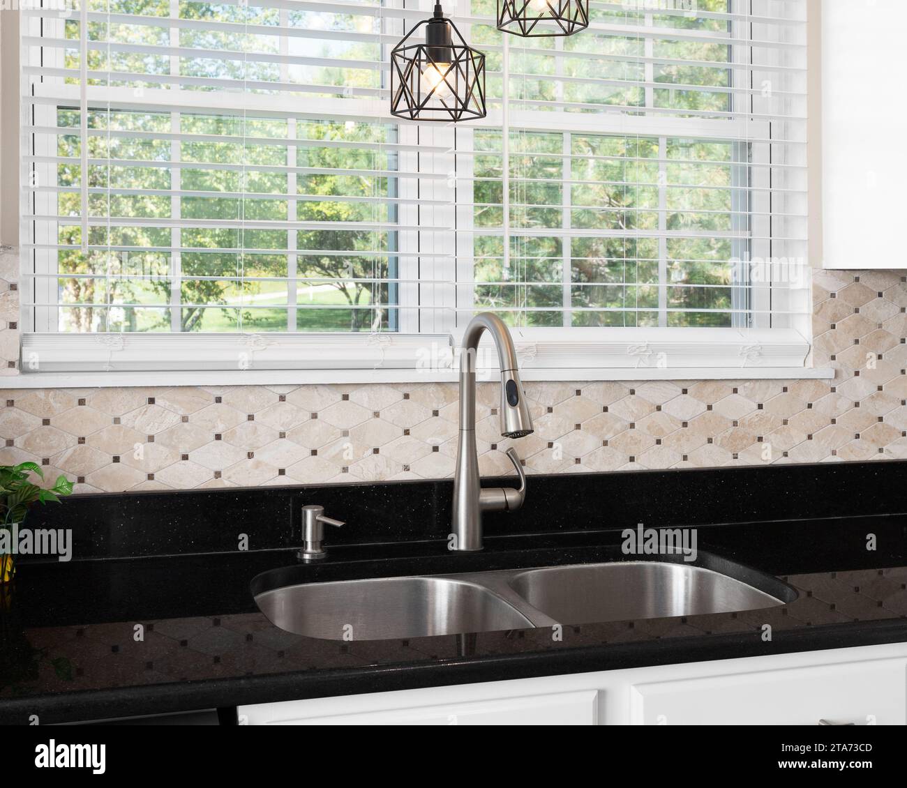 Un détail d'évier de cuisine avec un comptoir de quartz noir, un dosseret carrelé en pierre brune et des lumières suspendues au-dessus d'un robinet en acier inoxydable poli. Banque D'Images