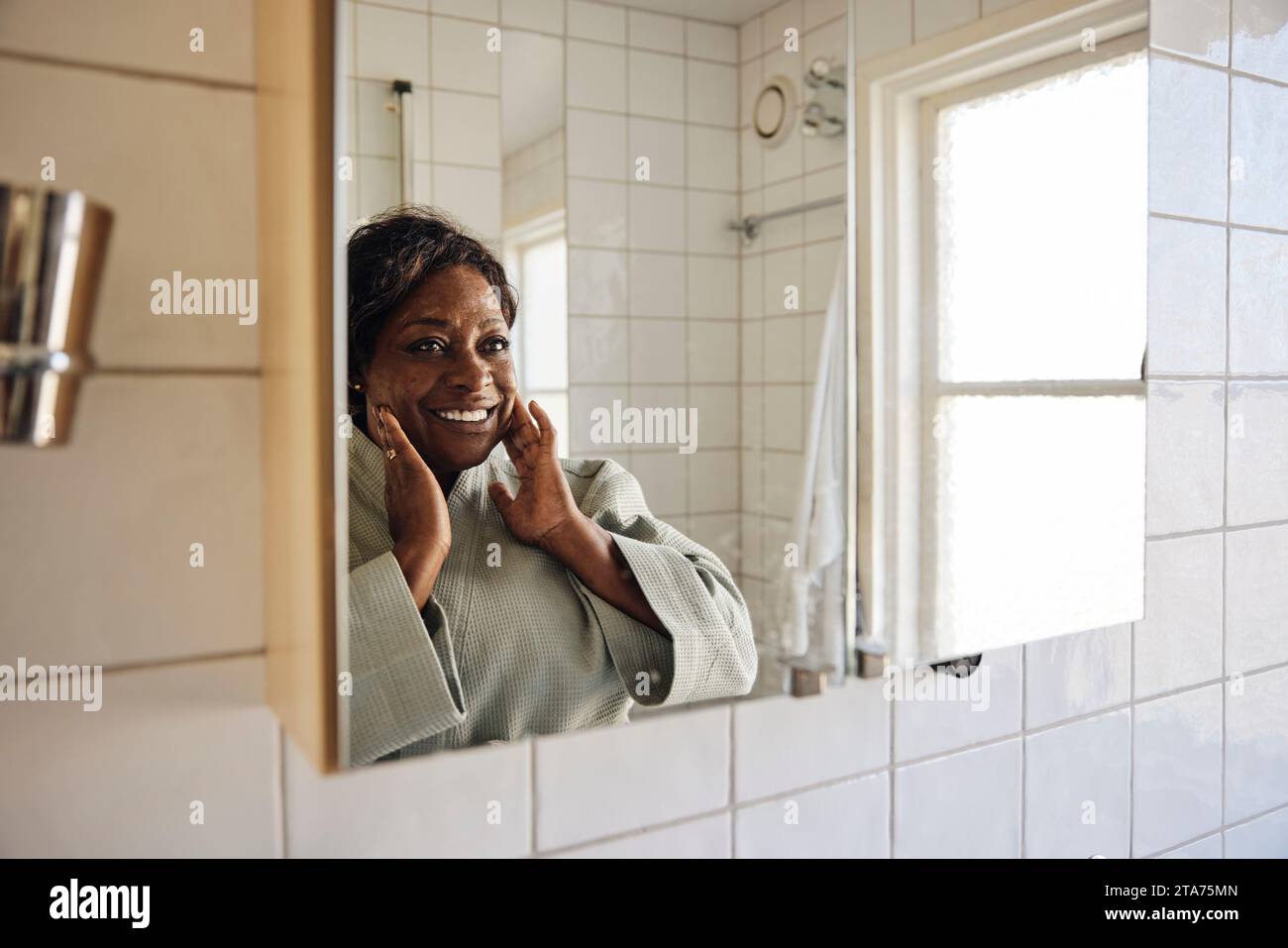 Heureuse femme mature touchant le visage tout en se regardant dans le miroir de salle de bain à la maison Banque D'Images
