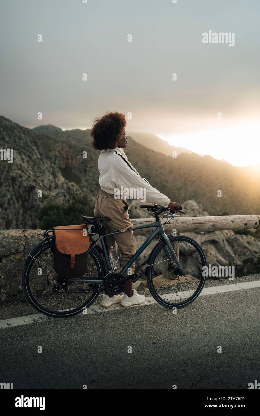 Femme explorant les montagnes tout en se tenant debout avec le vélo sur la route au coucher du soleil Banque D'Images