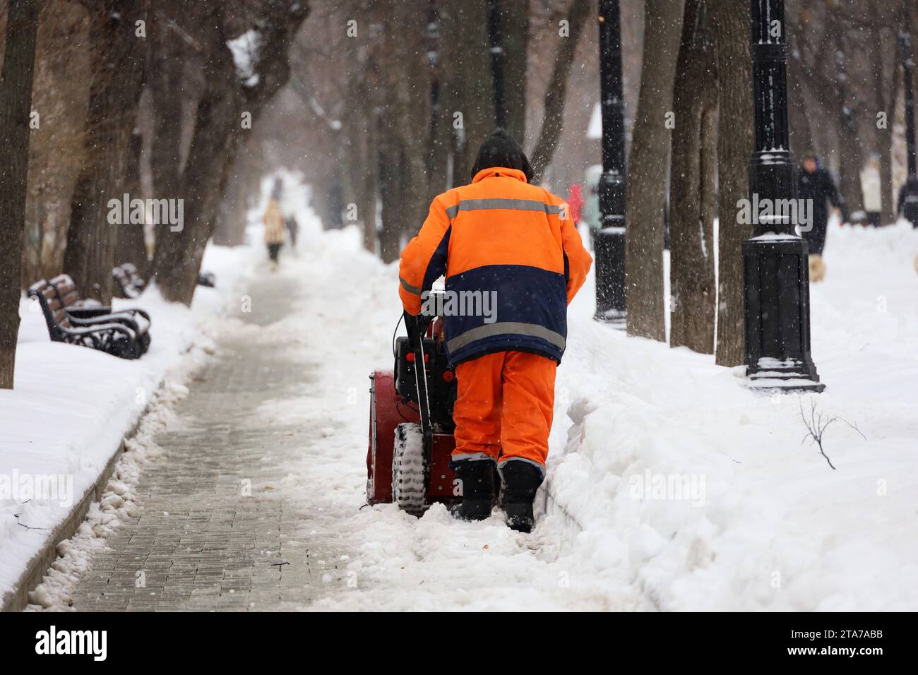 Travailleur des services communautaires en uniforme avec un chasse-neige déneigement sur un trottoir. Homme pendant le déneigement dans la ville d'hiver, nettoyage des rues Banque D'Images
