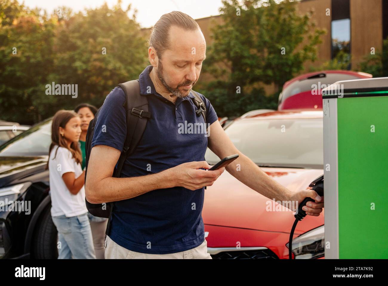 Homme utilisant le téléphone intelligent tout en tenant la fiche à la station de charge de voiture Banque D'Images