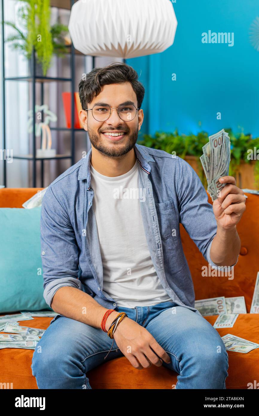 Gagnant heureux heureux gagnant Indien comptant l'argent acclamant avec des profits élevés, gain de jeu de loterie, satisfait des revenus de richesse salaire sur le canapé à la maison plein de billets de banque en dollars. Vertical Banque D'Images