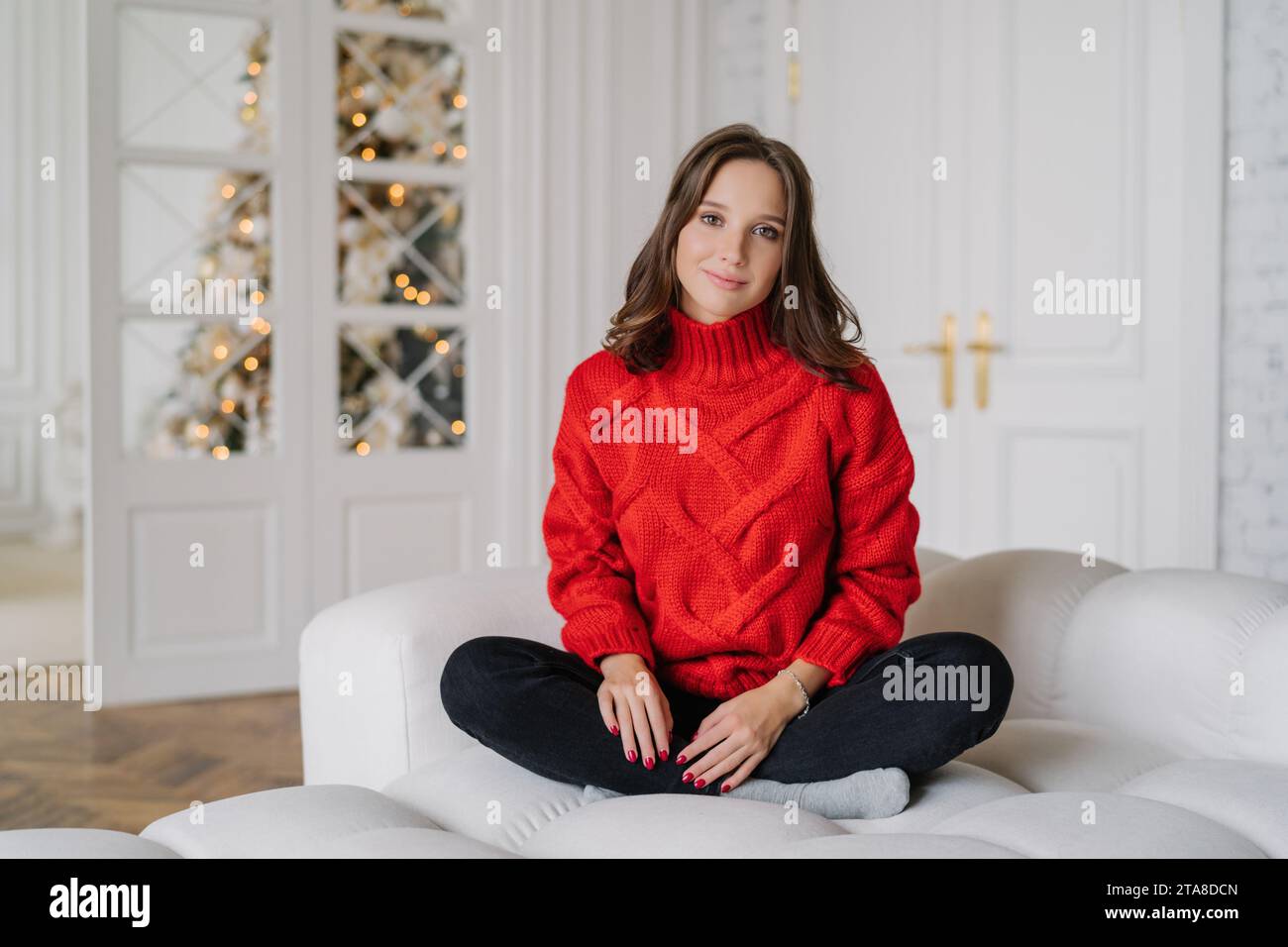 Jeune femme détendue dans un pull en tricot rouge vibrant assis sur un canapé, lumières festives en arrière-plan, incarnant l'esprit de vacances. Banque D'Images